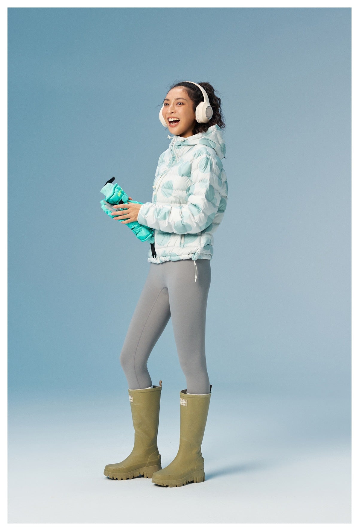 Side view of a model wearing a white quilted hooded jacket with aqua circular prints, gray leggings, and olive rain boots.