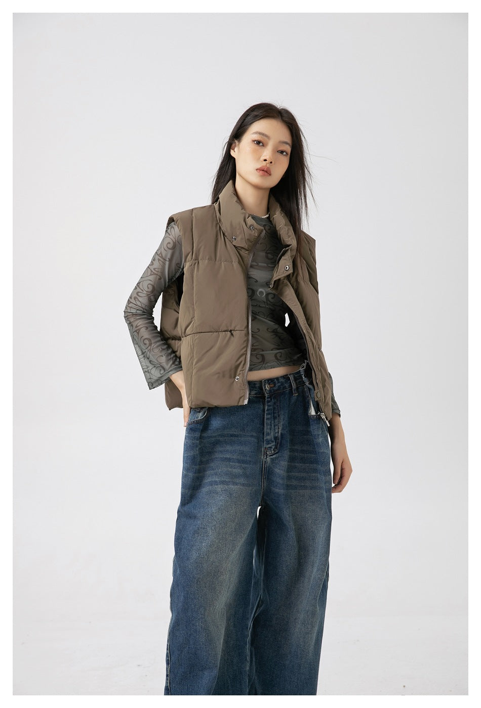 Model in a taupe padded vest over a gray patterned sheer top and wide-leg blue jeans, shown from head to mid-leg.