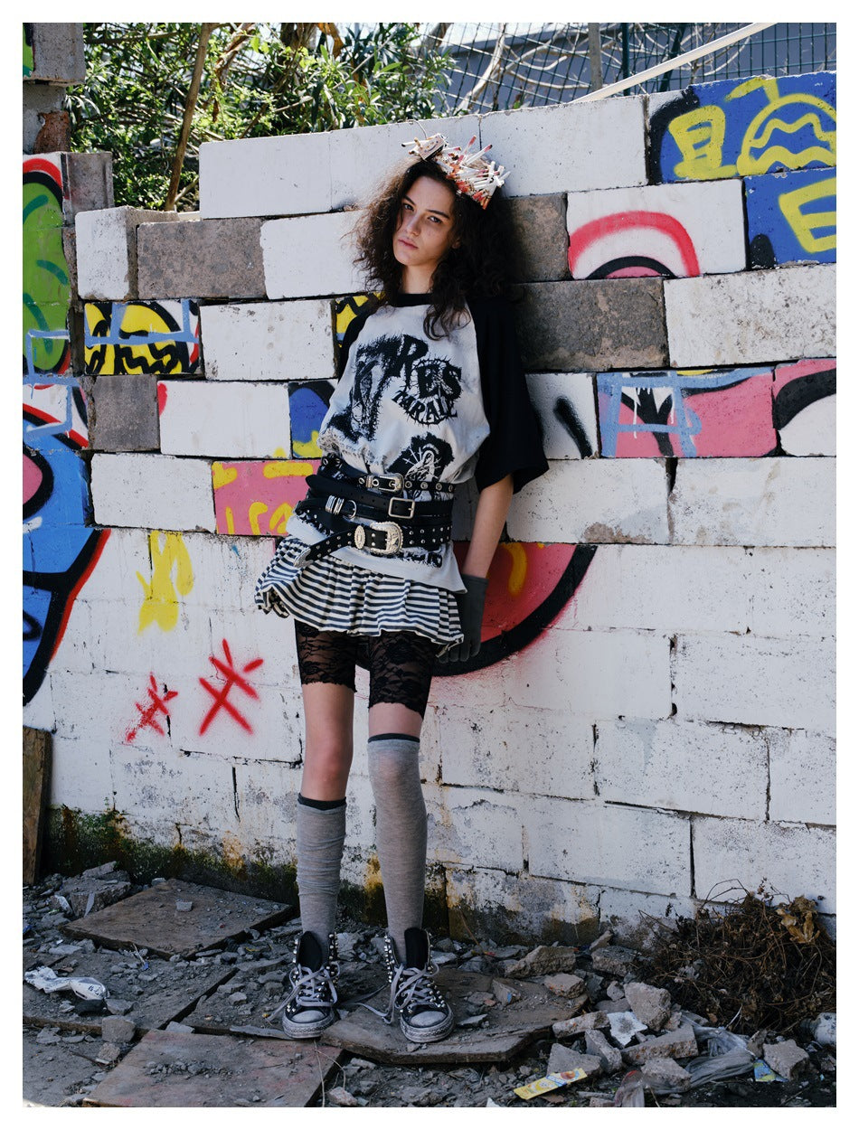 Model wears a black-and-gray graphic raglan shirt with a striped skirt and layered belts against a graffiti-covered wall.
