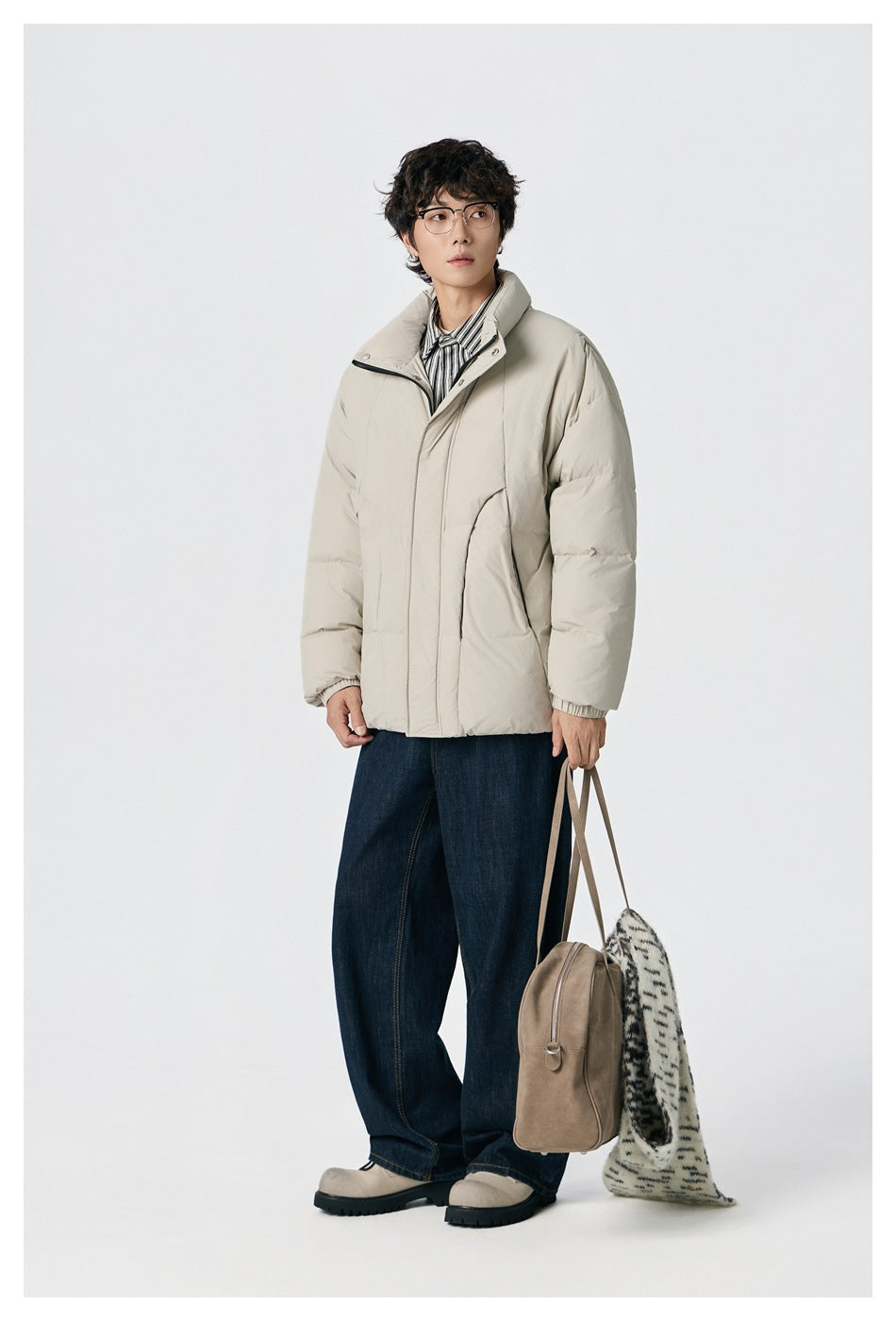 Man wearing beige puffer jacket over a striped shirt with dark wide-leg jeans, holding a tan bag