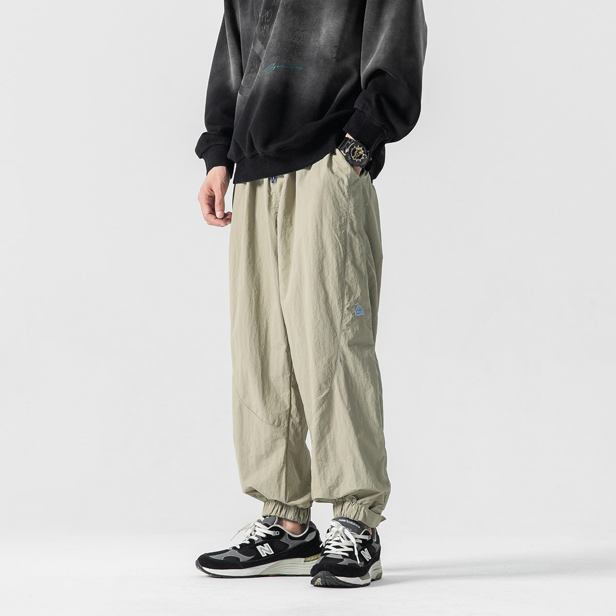 Model wearing light khaki jogger pants with a small blue emblem and elastic cuffs, shown from the front.