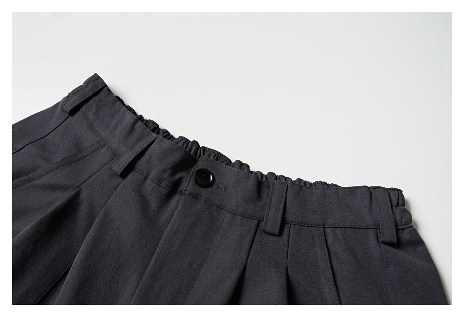 Close-up of charcoal trousers showing a buttoned waistband, belt loops, pleats, and elastic gathering at the back.
