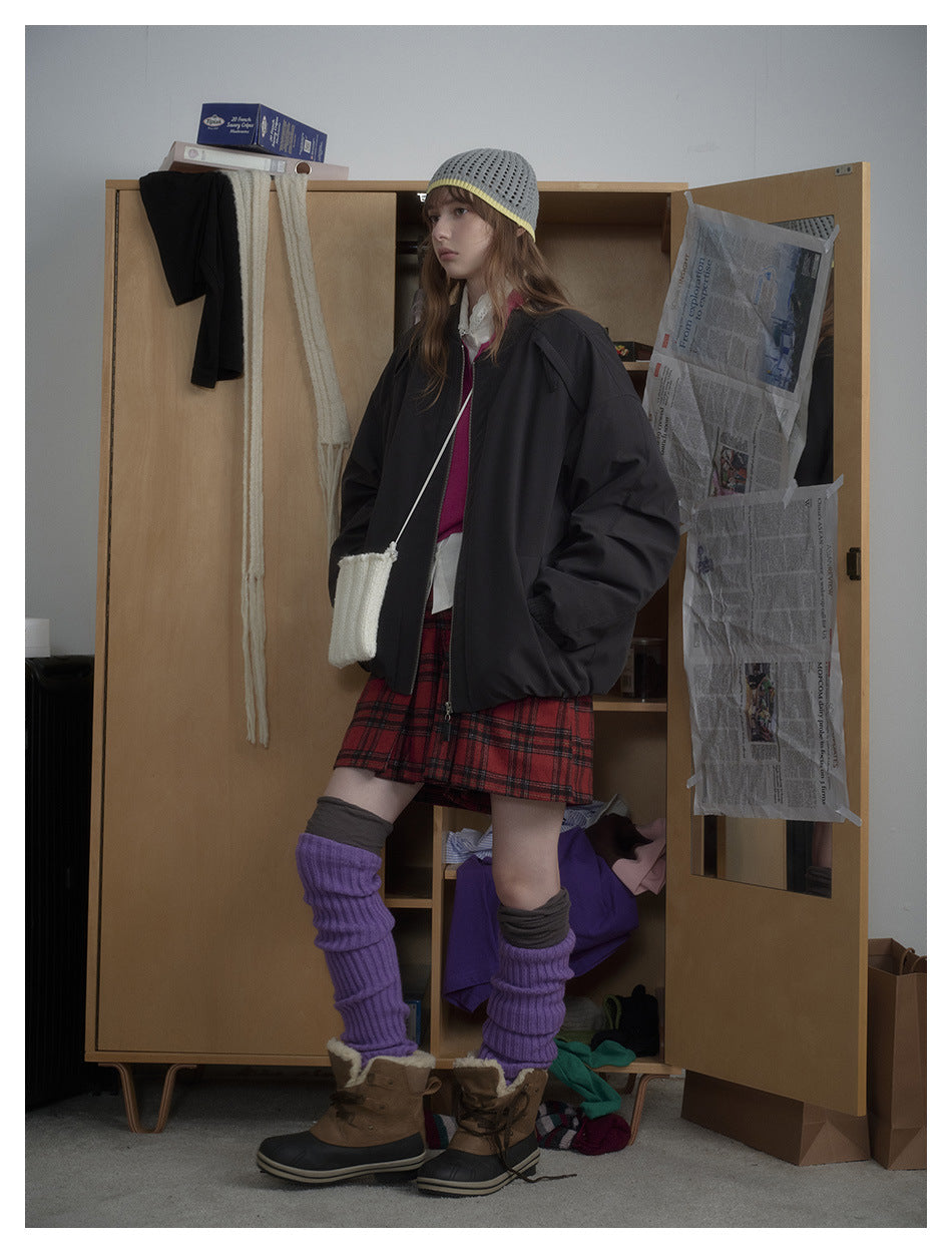 Model wearing the black zip bomber jacket in a three-quarter side view with a red plaid skirt and purple leg warmers