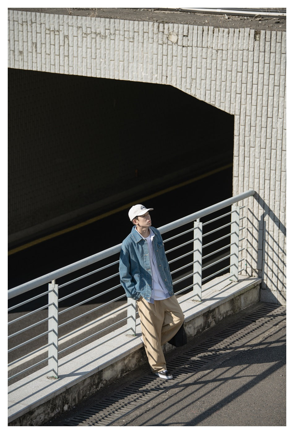 Distant model leaning by a railing, wearing a blue denim jacket, striped shirt, tan trousers, and white cap.