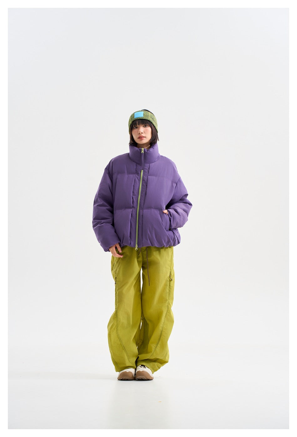 Model wearing a purple high-collar puffer jacket with lime zipper, loose lime trousers, and a green knit headband.