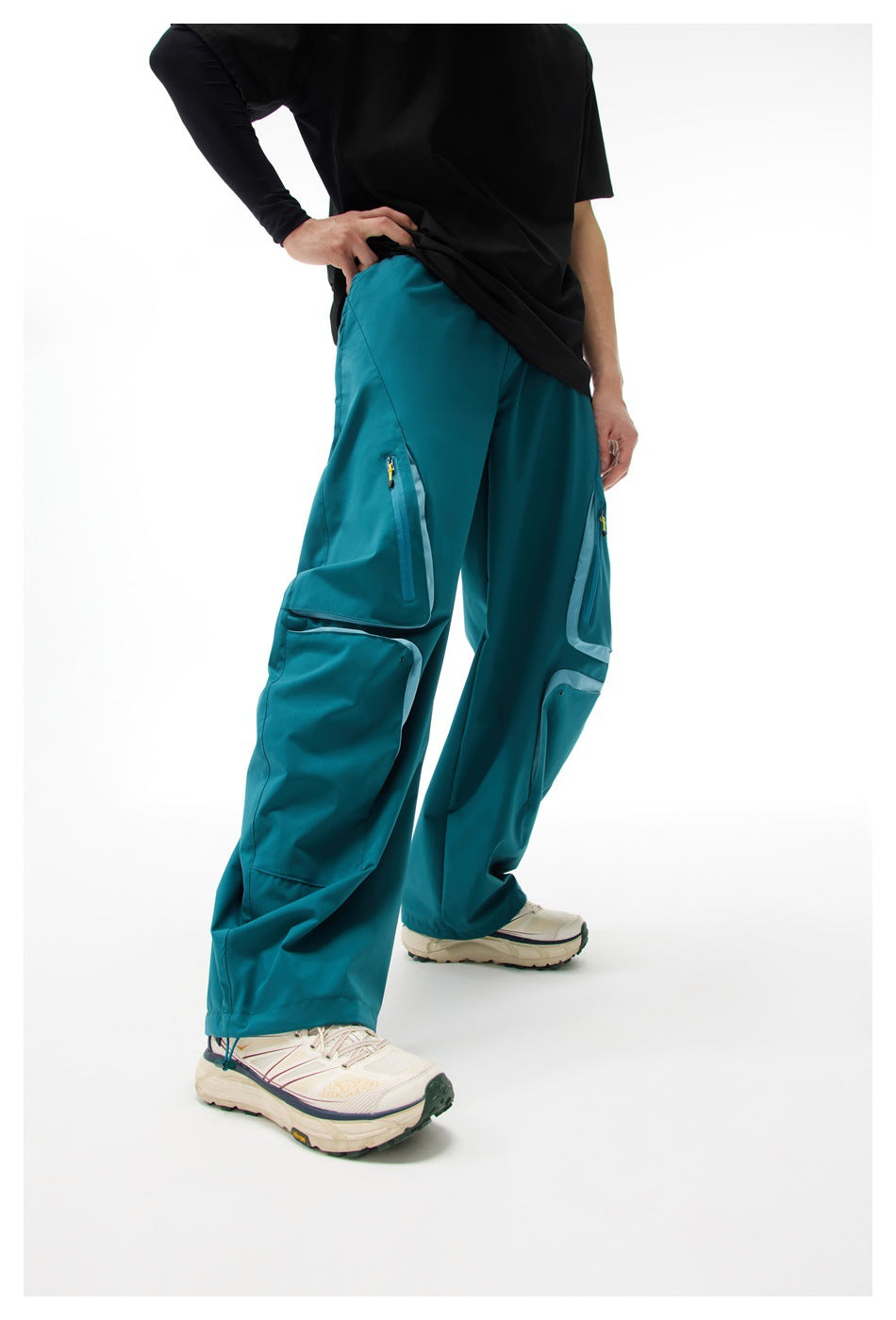Angled view of a model wearing teal cargo pants with large flap pockets and yellow-accented zippers.
