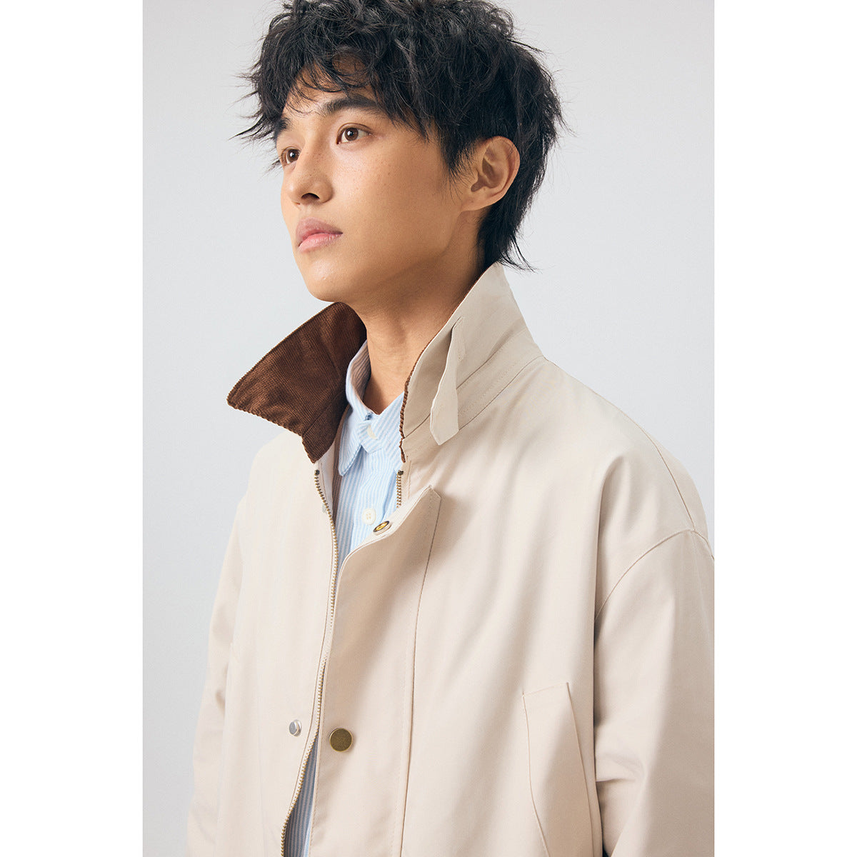 Close-up of a model wearing a beige coat, showing its raised brown collar, zipper, and snap-button placket.