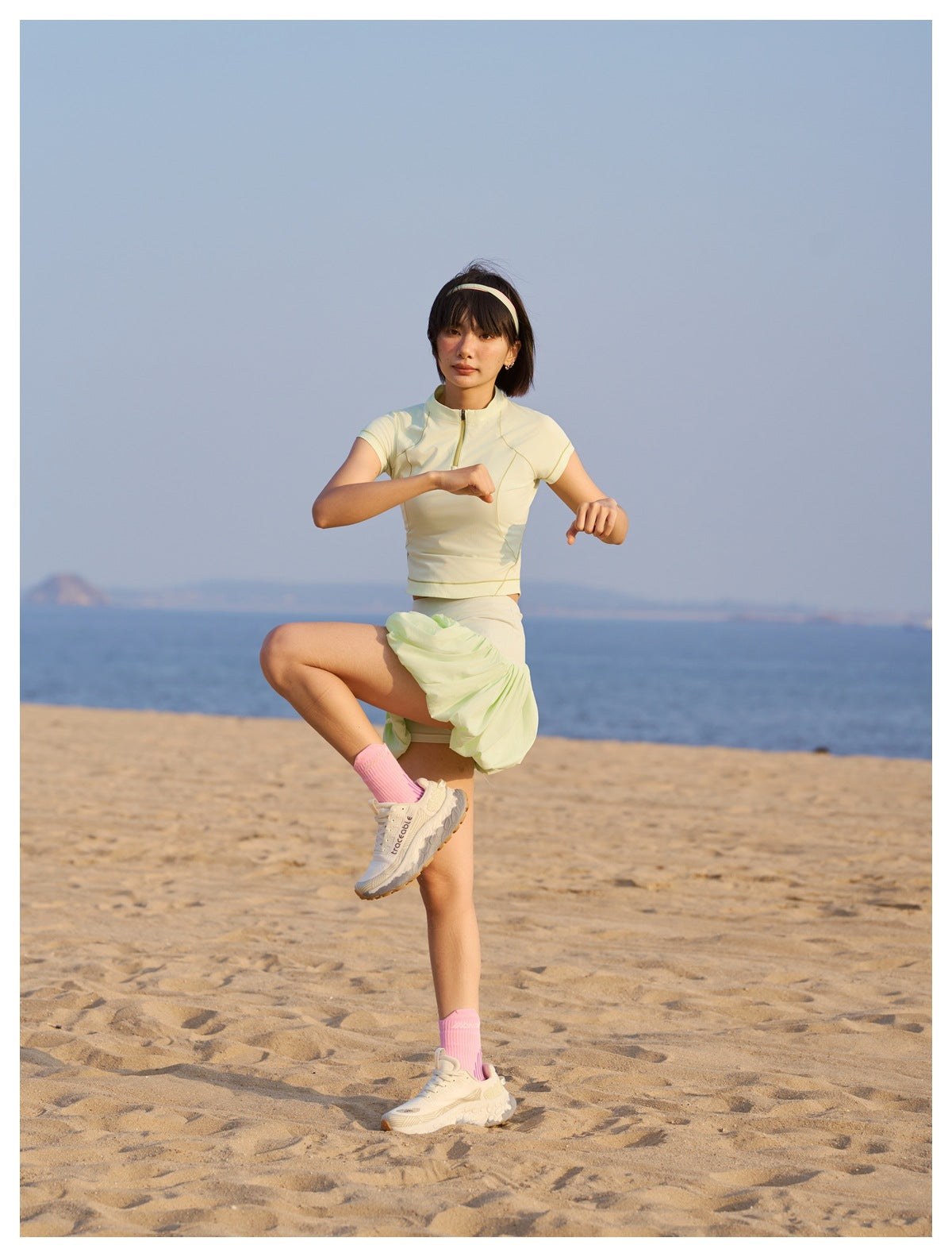 Model wearing a pale yellow short-sleeve zip-neck top and light green skirt on a beach.