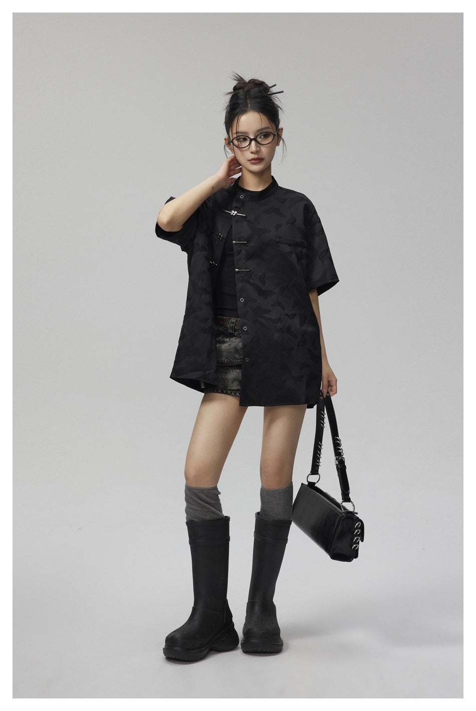 Model wearing an oversized black butterfly-pattern shirt with silver bar closures, styled with shorts and tall boots.