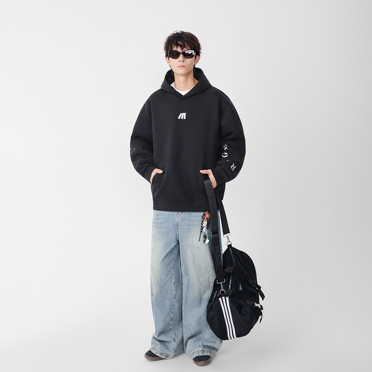Model wearing a black oversized hoodie with a white chest emblem and sleeve lettering, paired with wide-leg light-wash jeans.