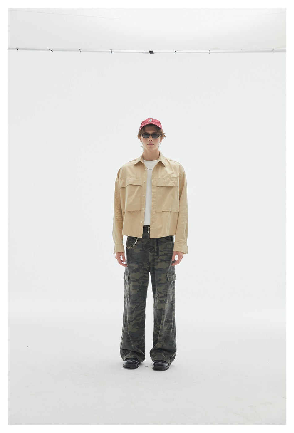 Model wearing a beige button-up overshirt with large front pockets and loose dark camouflage cargo pants.