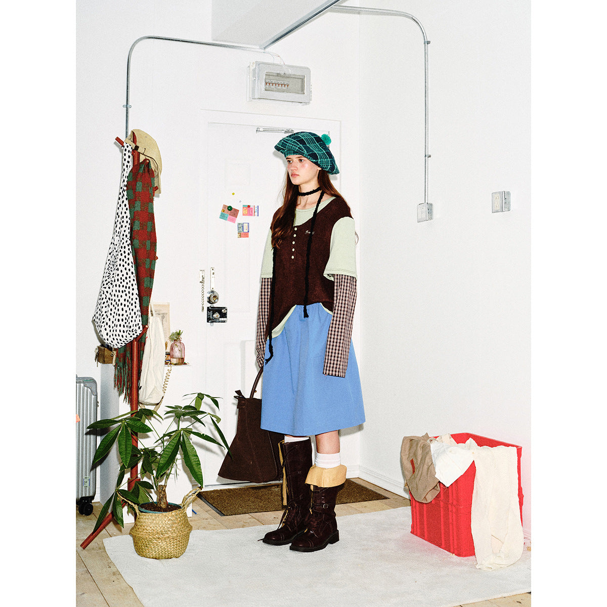 Model in a green plaid beret, layered sage and burgundy top, blue skirt, and dark boots, shown in a room.