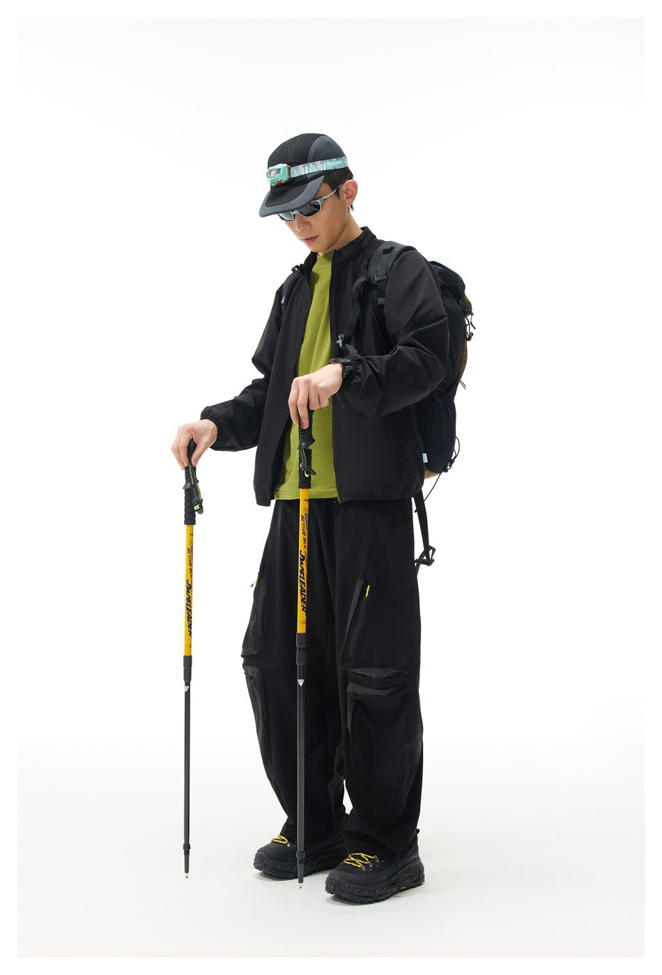 Model wearing a black zip jacket over a green shirt with black cargo pants, backpack, and hiking poles.