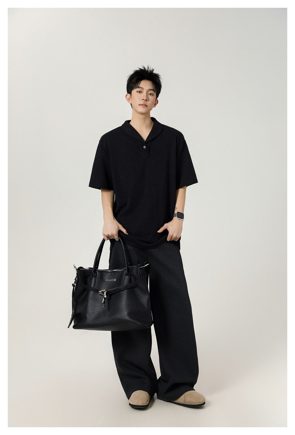 Full-length view of a model in a black textured polo and dark wide-leg trousers, carrying a black tote.
