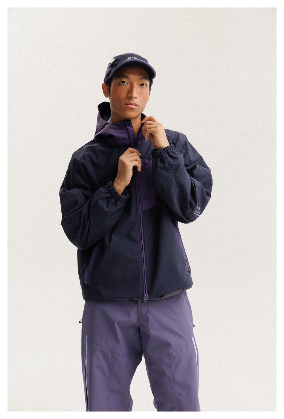 Model adjusts the collar of a navy and purple hooded jacket, paired with purple pants.