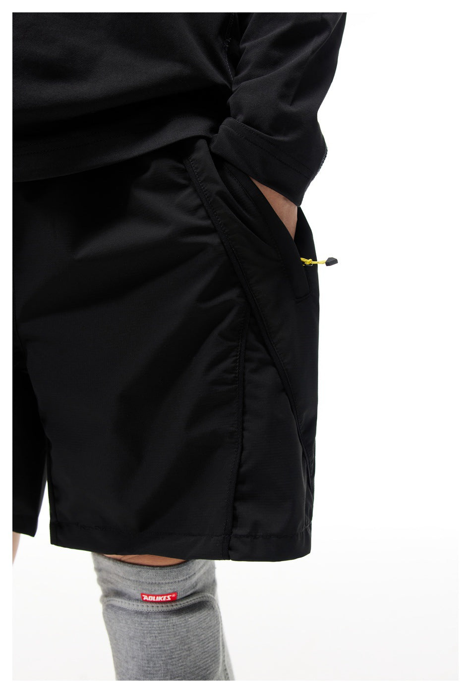 Close-up of black shorts with a hand in the diagonal side pocket showing seam detail and a yellow zipper pull cord