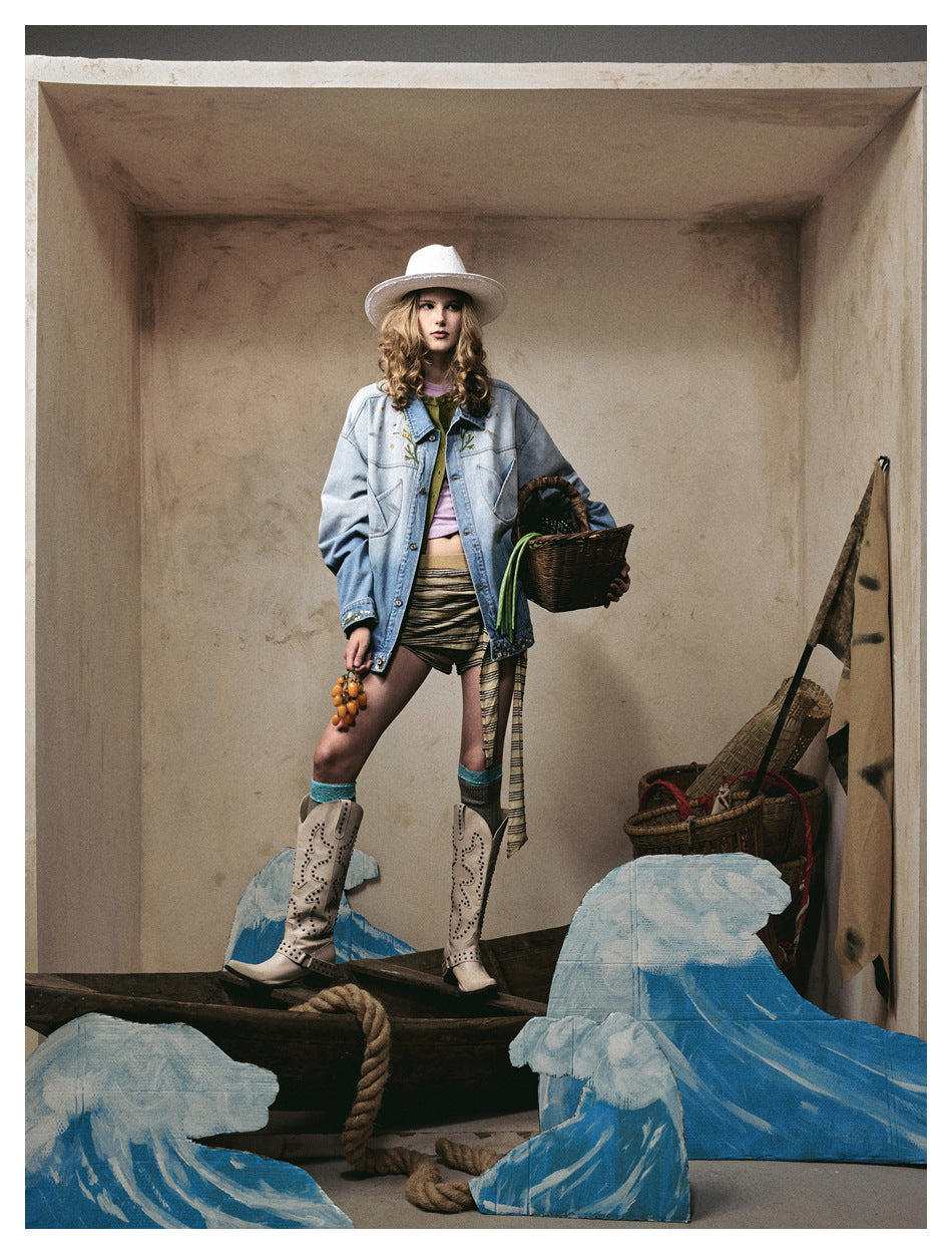 Model standing in a styled set wearing an open oversized blue denim jacket with a hat, shorts and boots