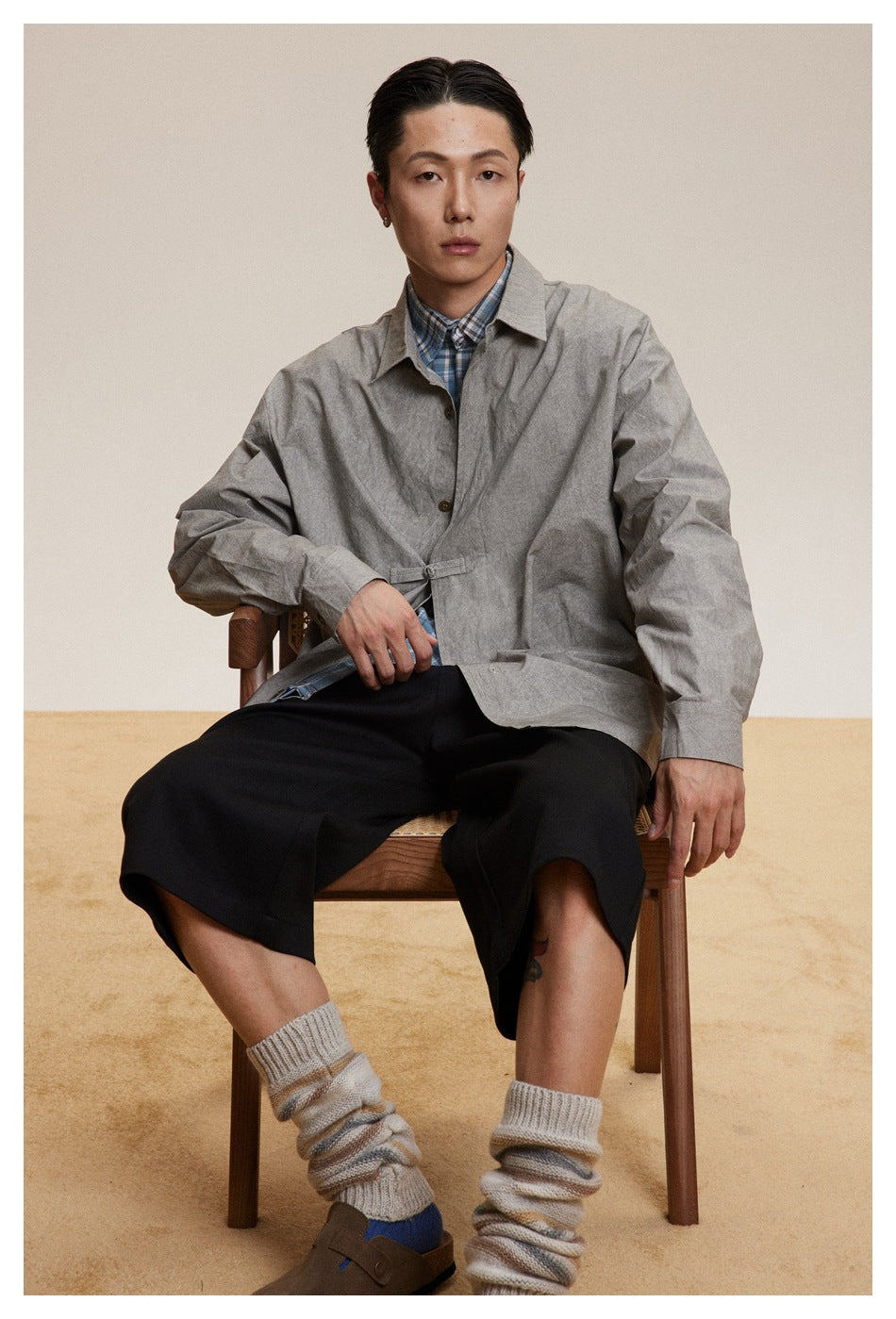 Model seated wearing a light gray button-front jacket with a small waist tab over a blue plaid shirt.