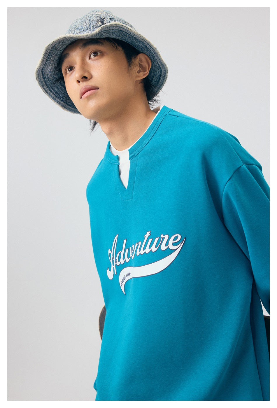 Close-up of a model wearing a bright teal sweatshirt with a notched neckline and white Adventure chest graphic.
