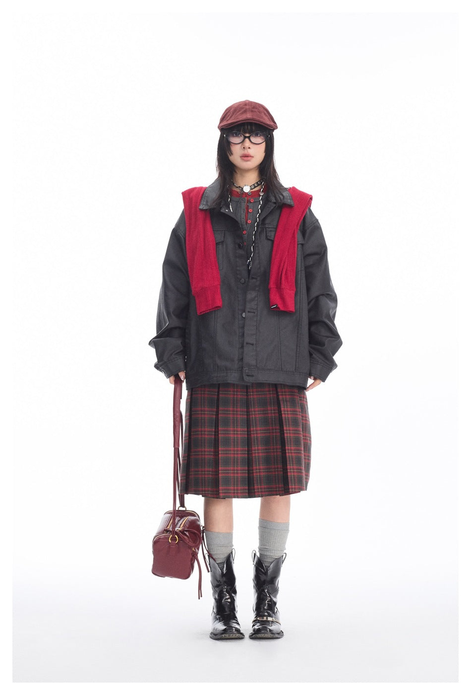 Model wearing oversized black coated jacket with red sweater over shoulders, plaid pleated skirt and black boots