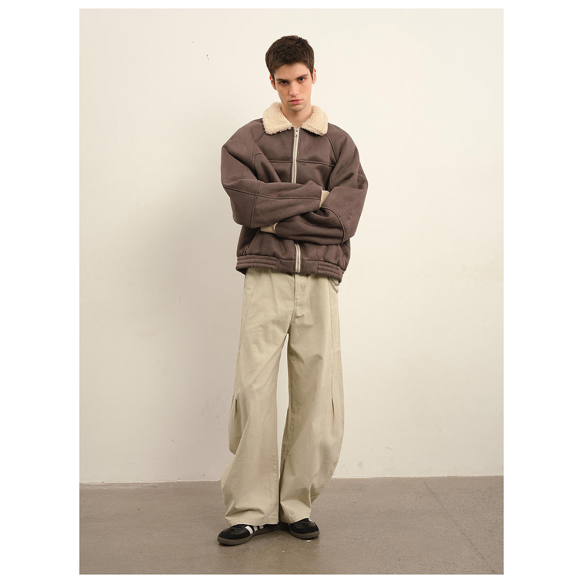 Model wearing a dark taupe jacket with a cream shearling-style collar and zip front, paired with loose beige trousers.