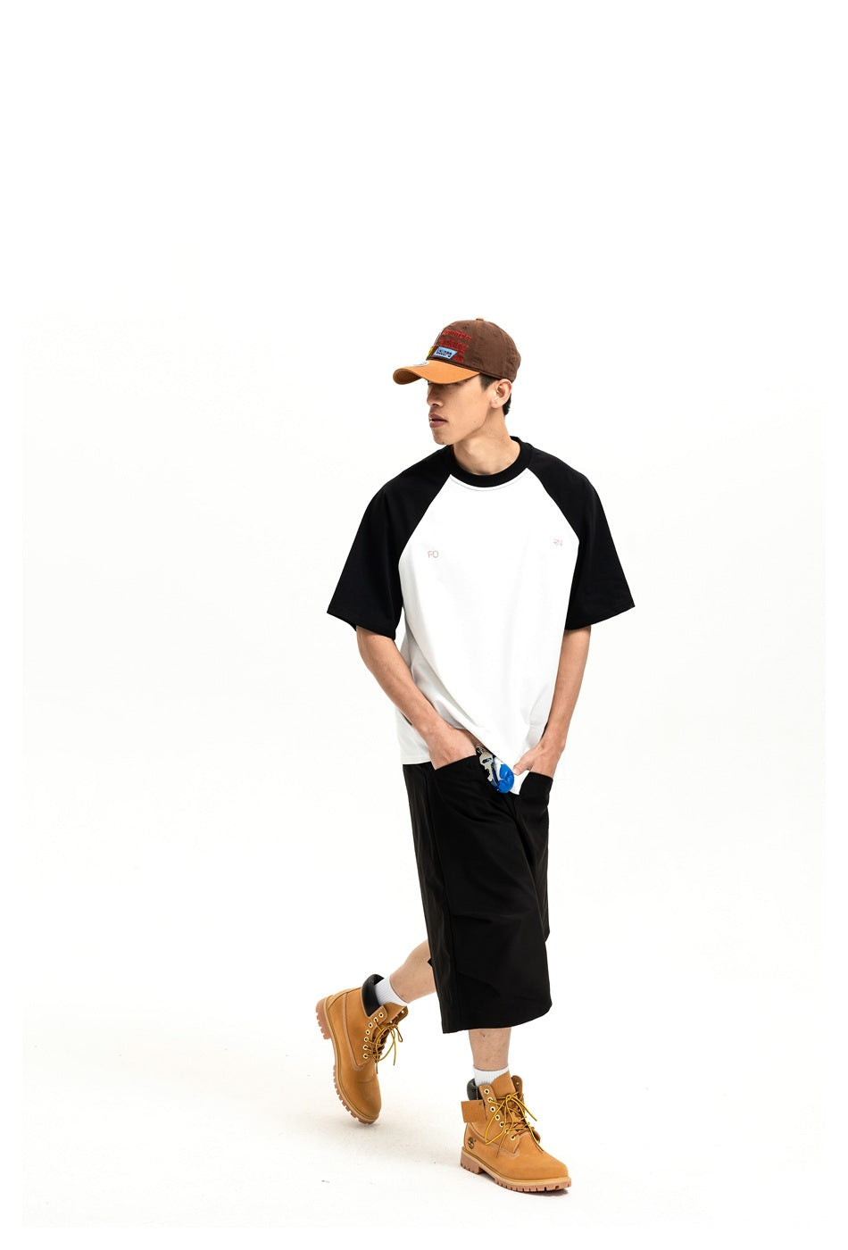 Model wearing a white raglan T-shirt with black sleeves and small red chest lettering, with black shorts, a cap and boots