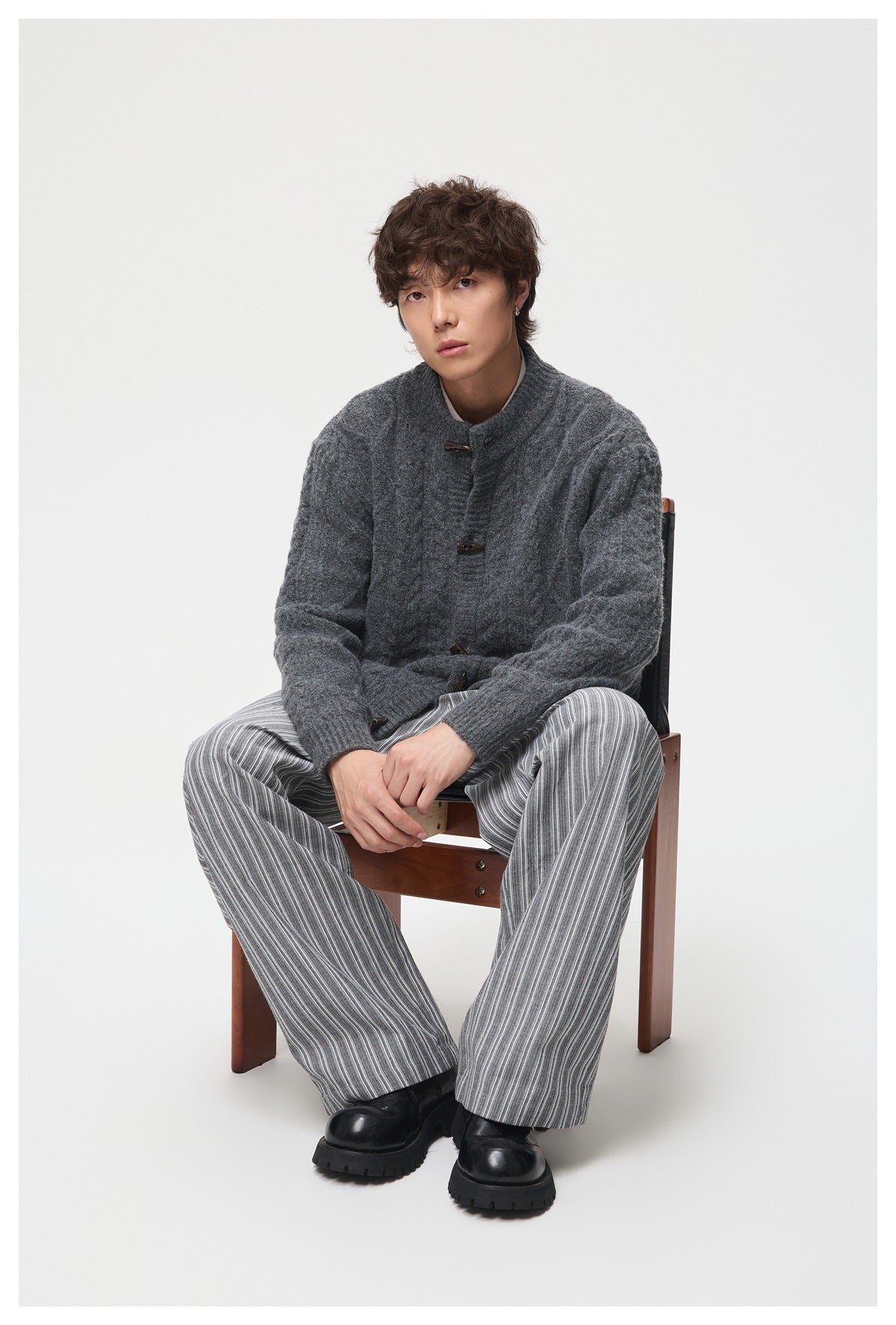Man seated on a wooden chair wearing a dark gray cable-knit toggle cardigan with striped wide-leg pants