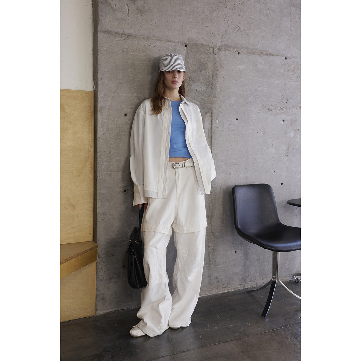 Model wears a white oversized button-up shirt and loose white trousers over a blue top, with a striped cap.