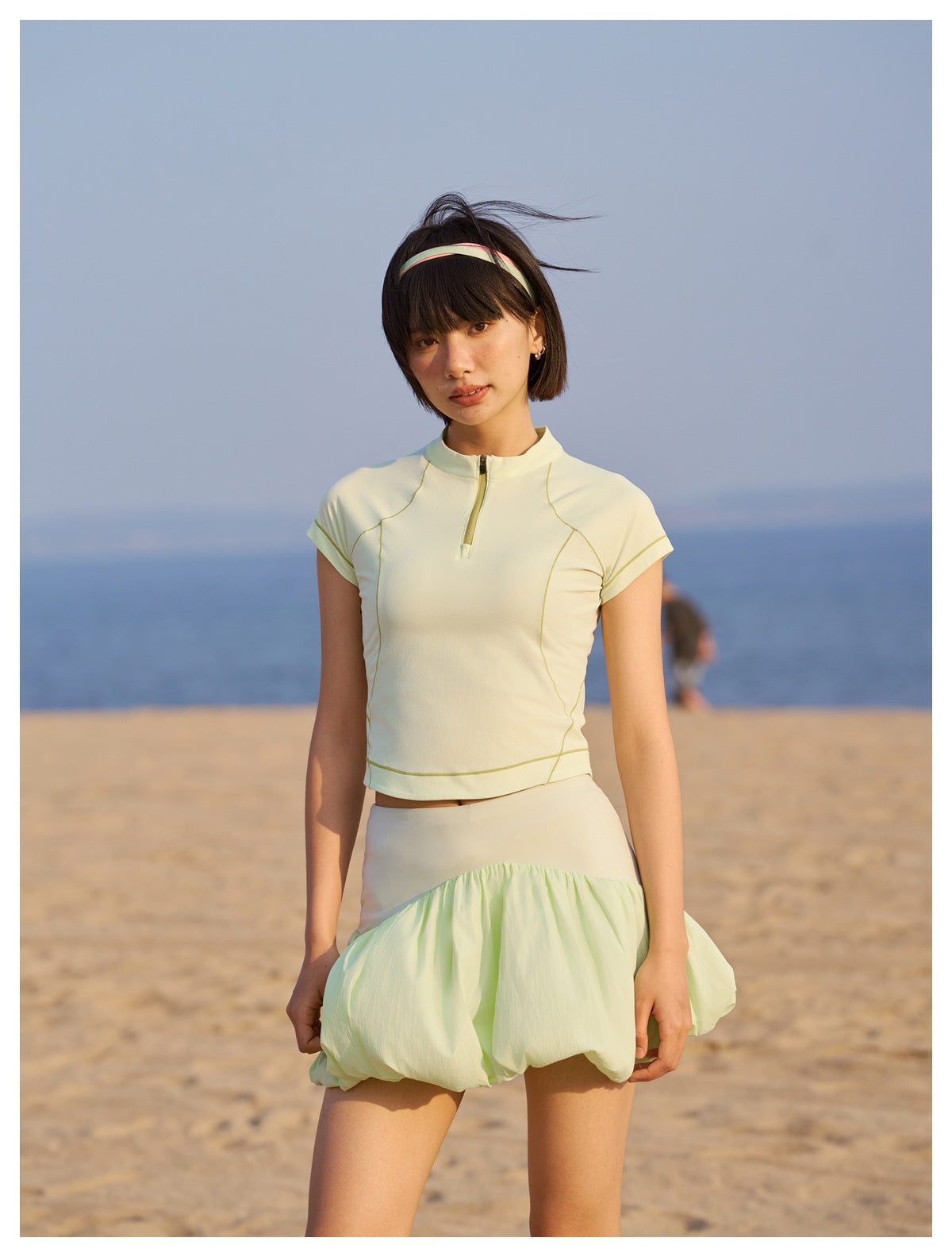 Model on a beach wearing a pale yellow-green zip-neck top and bubble skirt, shown from head to mid-thigh.