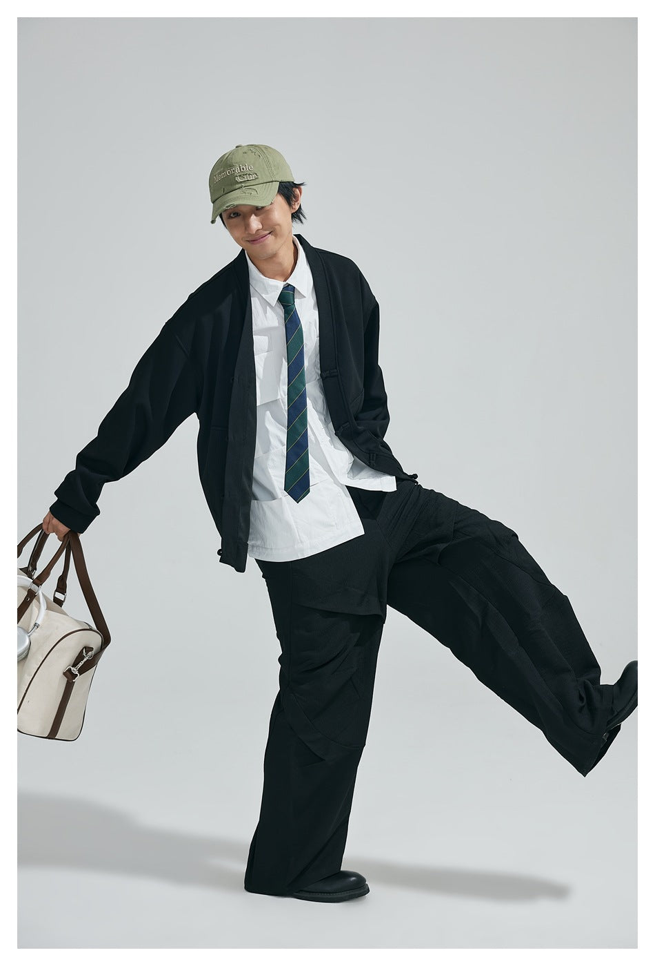 Model in a black cardigan and wide-leg trousers, posing with one leg raised and holding a duffel bag.