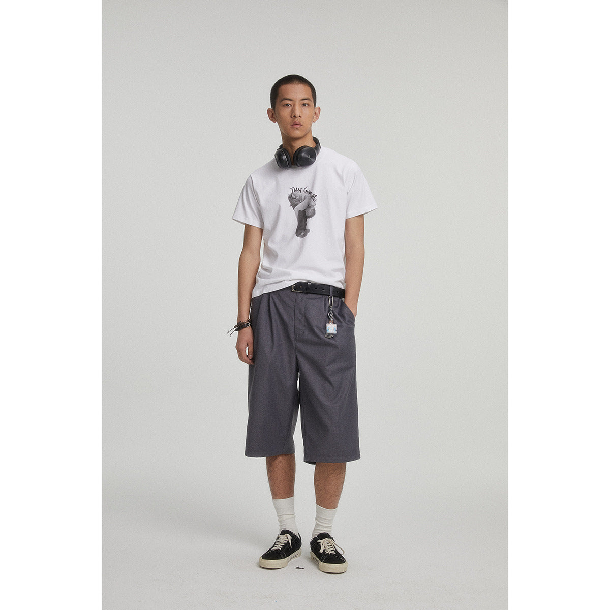 Model wearing a white graphic T-shirt and loose charcoal shorts, with headphones around his neck.