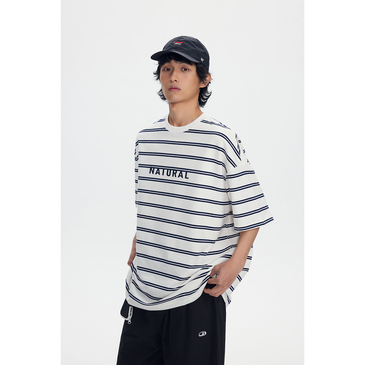 Model wearing an oversized ivory T-shirt with navy stripes and NATURAL lettering, shown from the front with black pants.