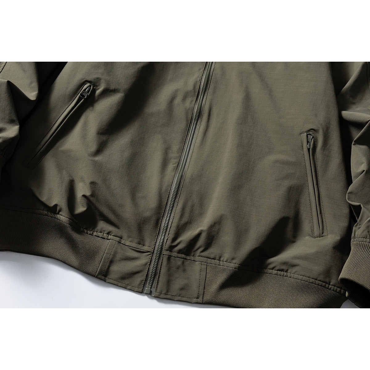 Close-up of an olive bomber jacket front showing two zip pockets, center zipper and ribbed hem and cuffs.