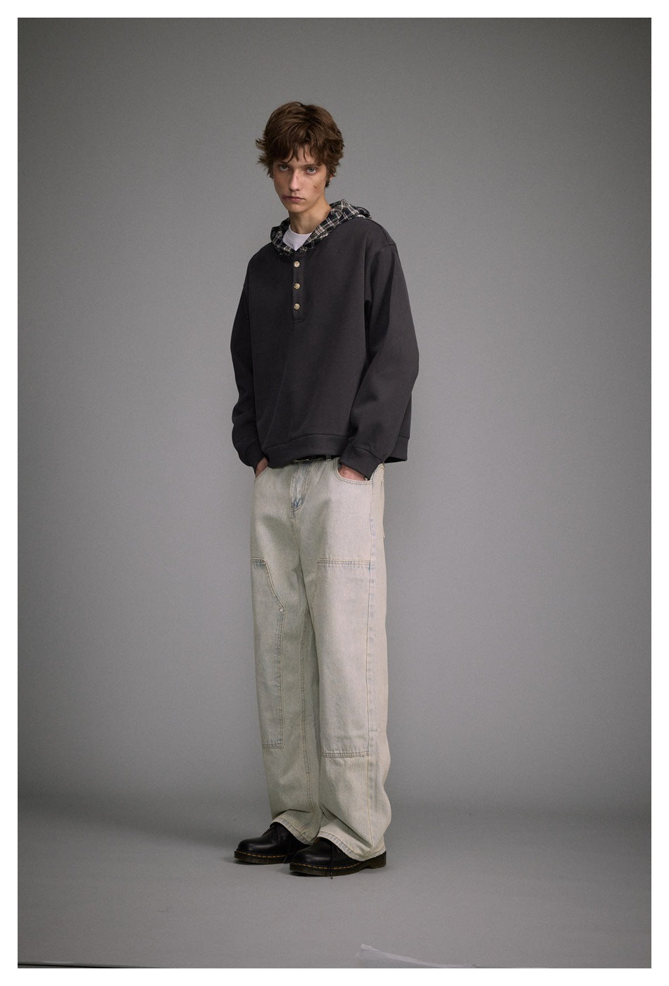 Model in a dark gray plaid-hood henley hoodie with hands in pockets of light washed jeans, three-quarter view