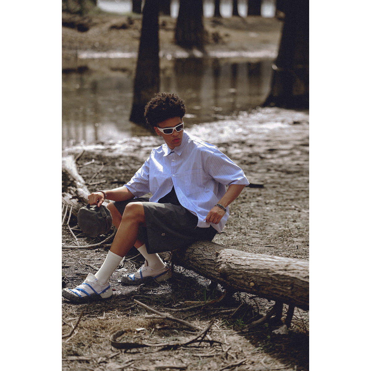 Model seated outdoors in an oversized pale blue pinstriped short-sleeve button-up shirt and dark shorts.