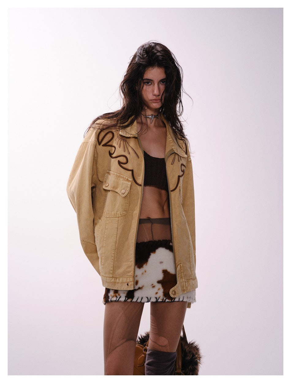 Model wearing an open tan jacket with dark embroidered motifs and flap pockets, shown with a black crop top and patterned