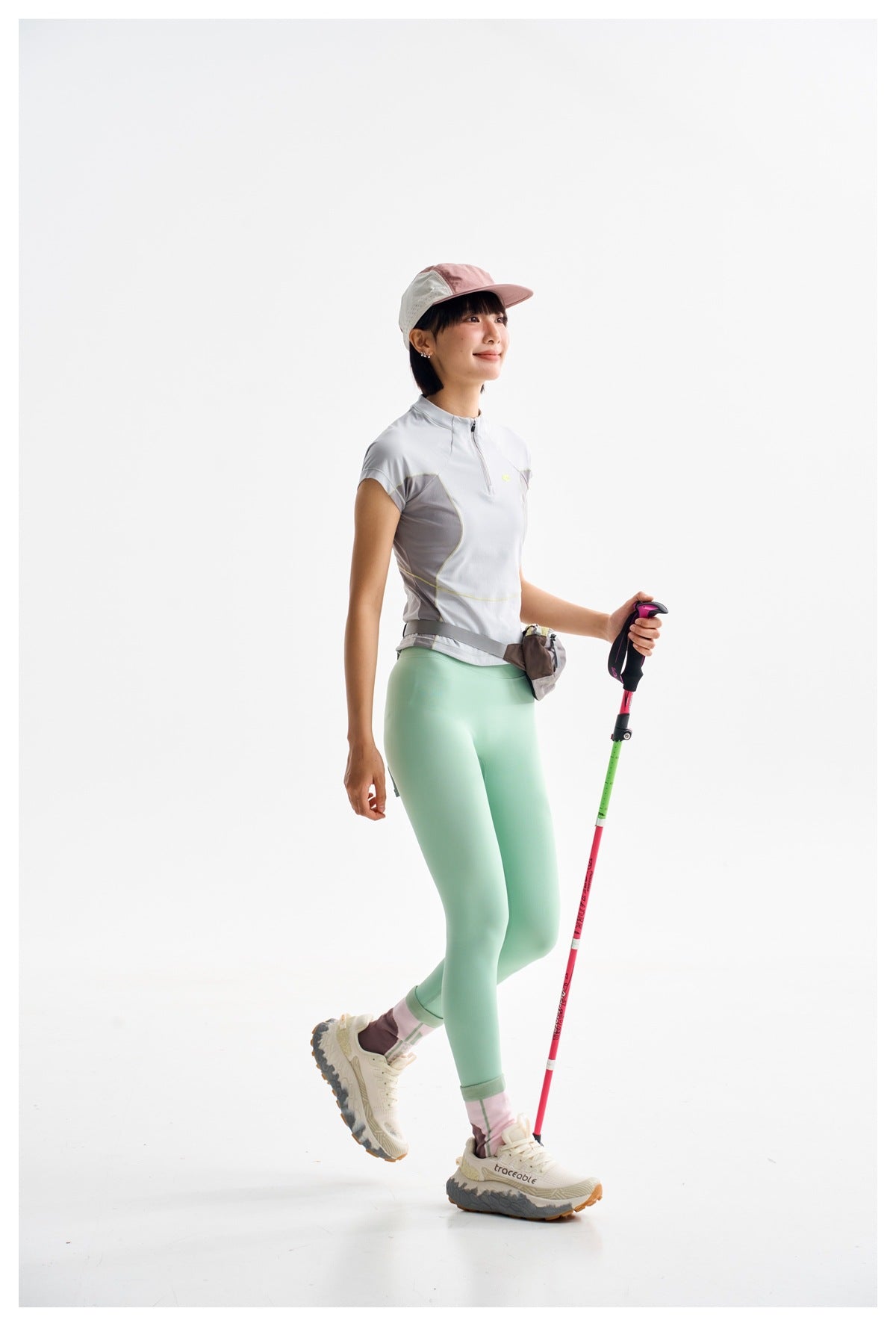 Woman in a white and gray short-sleeve quarter-zip top, mint leggings, and pink-and-white cap, holding a trekking pole.