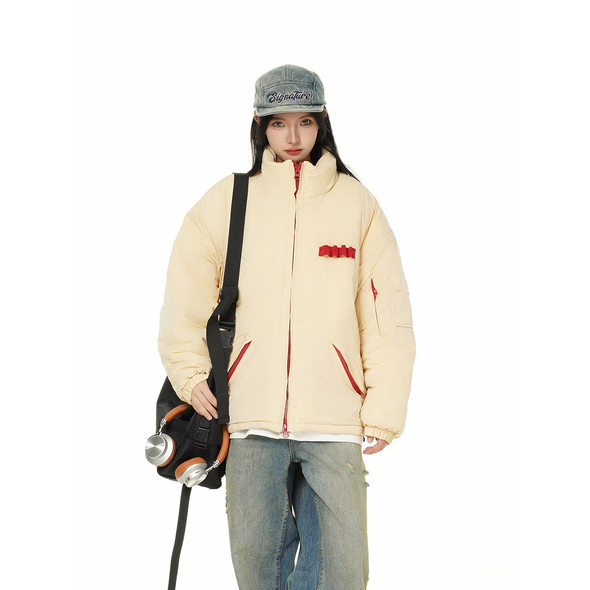 Model wearing a cream padded jacket with red details and wide jeans, carrying a black bag with headphones.