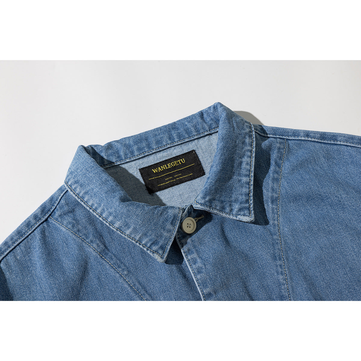 Close-up of a blue denim jacket collar, shoulder seams, button, and black woven neck label.