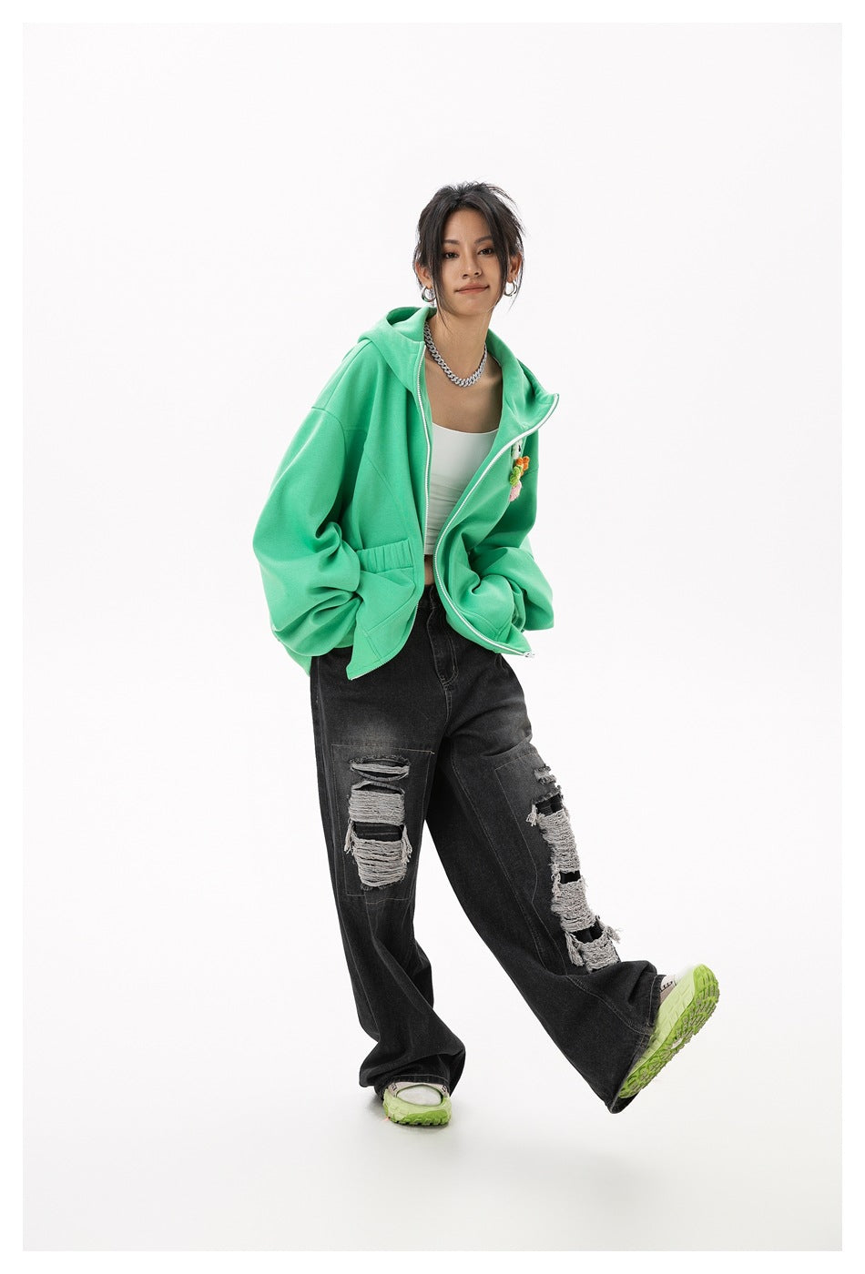 Model in a green zip hoodie with hands in pockets, white top, ripped black jeans and green sneakers
