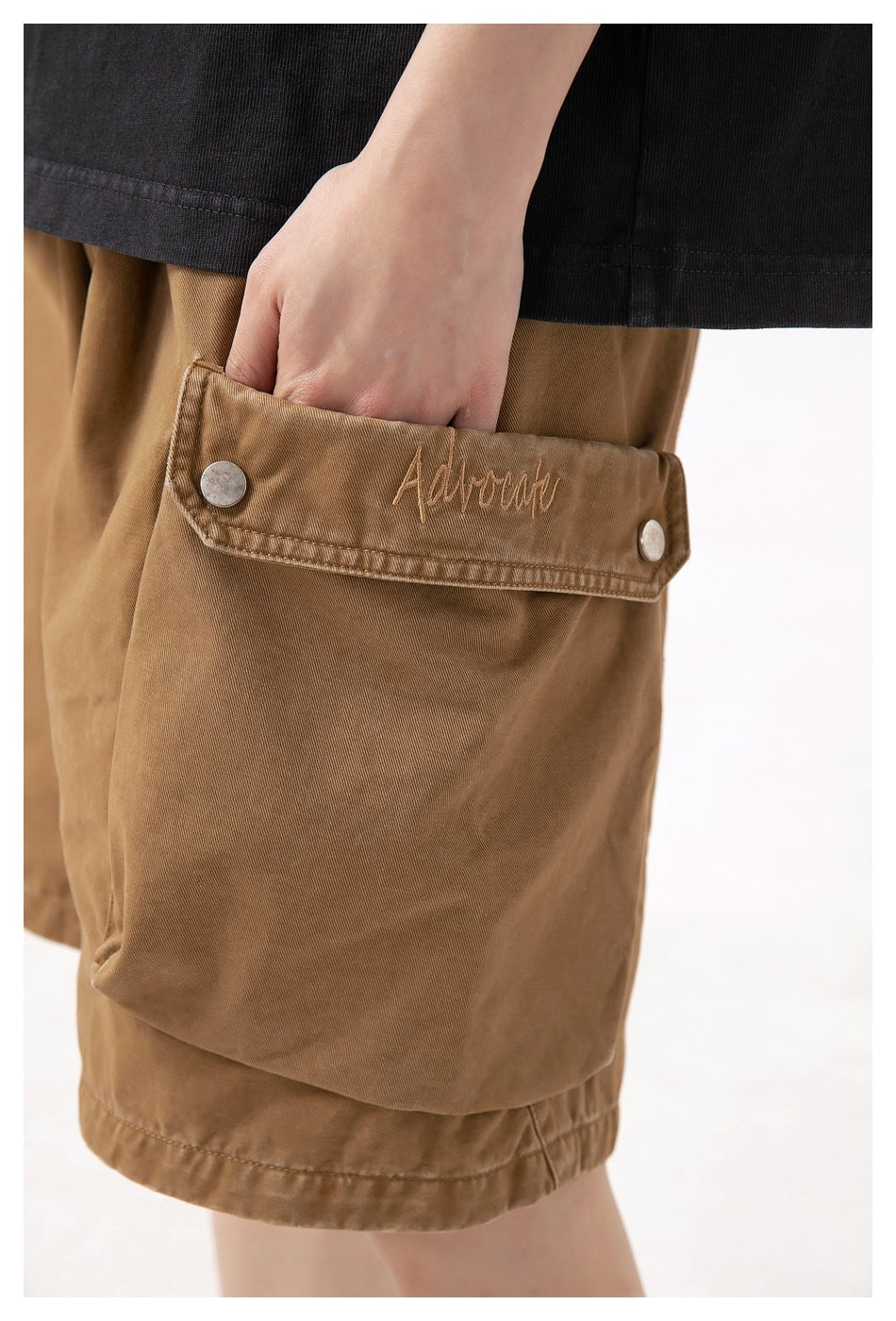 Close-up of brown cargo shorts showing a large flap pocket with metal snaps and embroidered script.