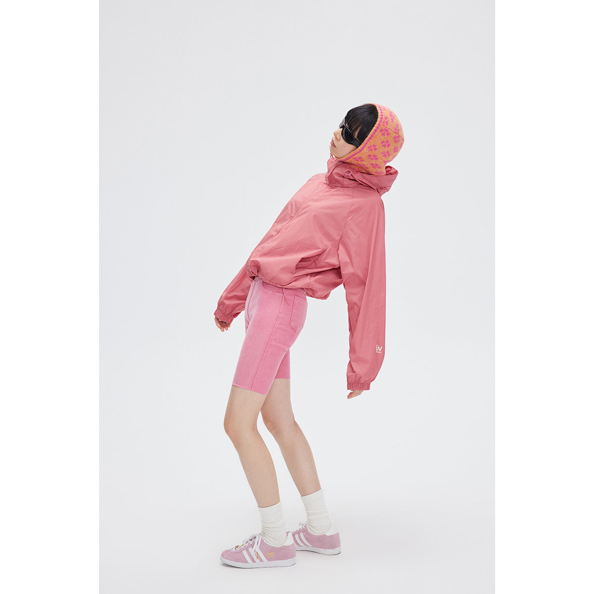 Model wearing a rose-pink hooded jacket and pink cycling shorts, leaning back in sunglasses and a patterned hood.