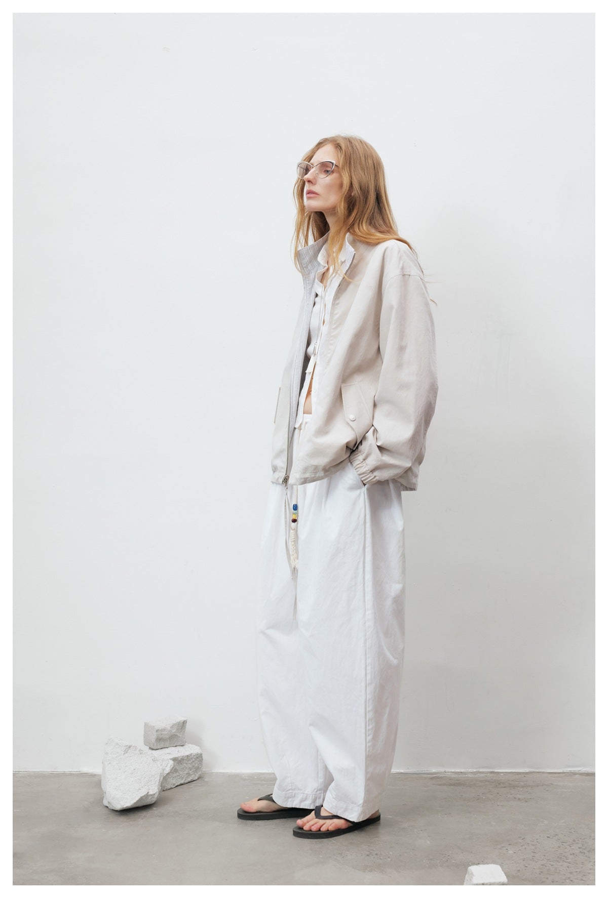 Model standing in profile wearing the beige zip jacket open over a white shirt and wide white pants