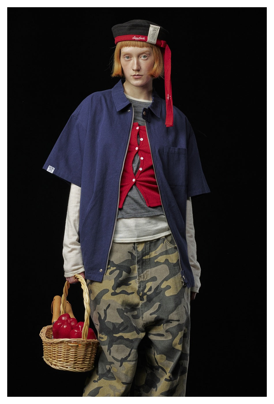 Model wearing an open navy short-sleeve zip shirt over layered tops with loose camouflage-print trousers.
