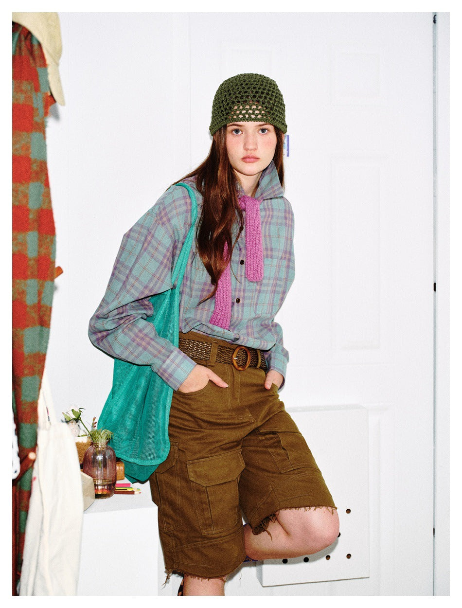 Model wearing the plaid shirt open at the collar with a pink knit tie, green beanie, olive cargo shorts and teal tote