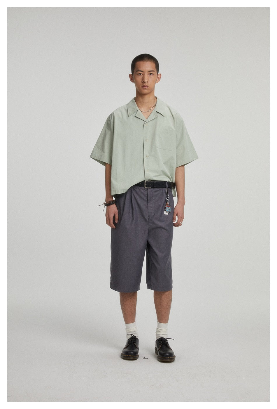 Front view of a model wearing a pale sage short-sleeve button-up shirt and loose charcoal shorts.