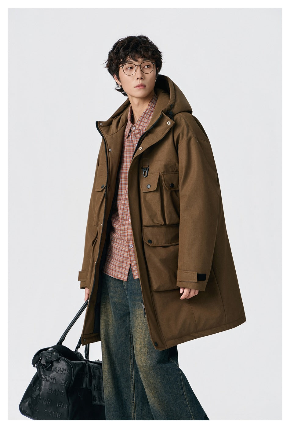 Model wearing a long brown hooded parka with multiple flap pockets, layered over a plaid shirt.