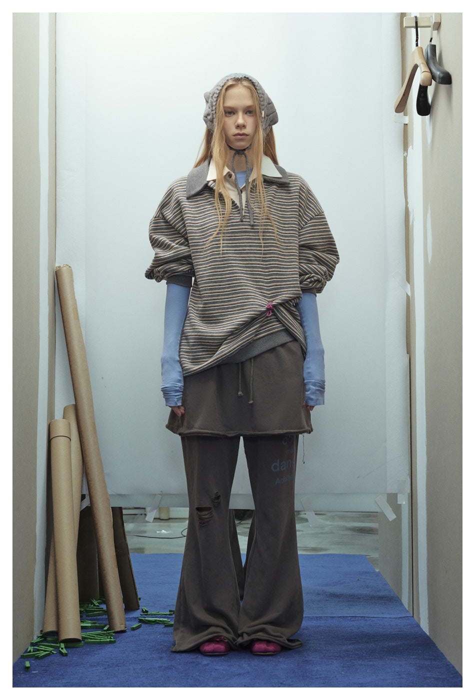 Full-length model wearing a loose gray striped polo over blue sleeves, with dark wide-leg pants and a gray hood.