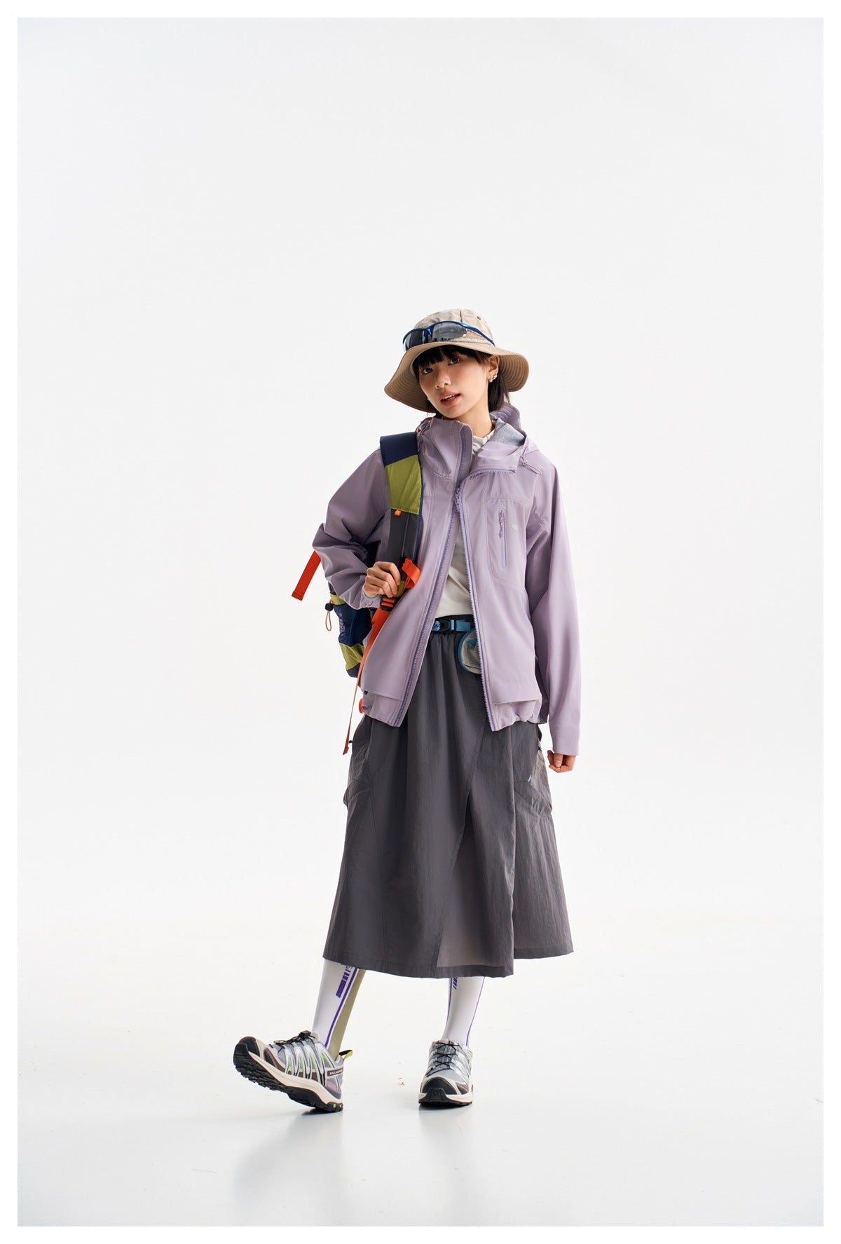 Model wearing a light lavender hooded jacket with zip pockets, dark gray skirt, and hiking accessories.