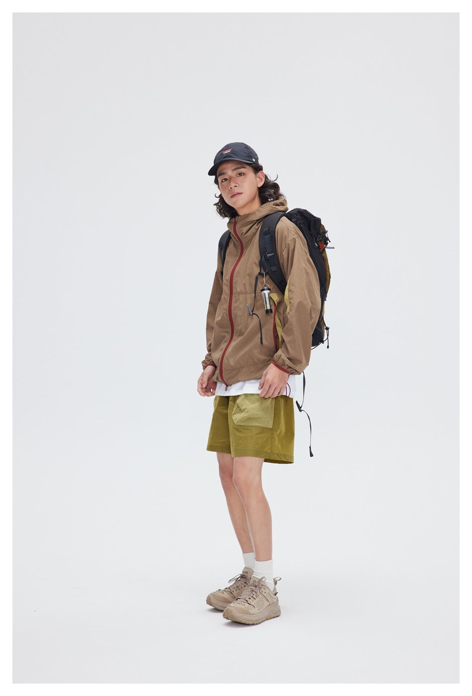 Three-quarter view of a model in a tan hooded jacket and olive shorts with a black backpack.